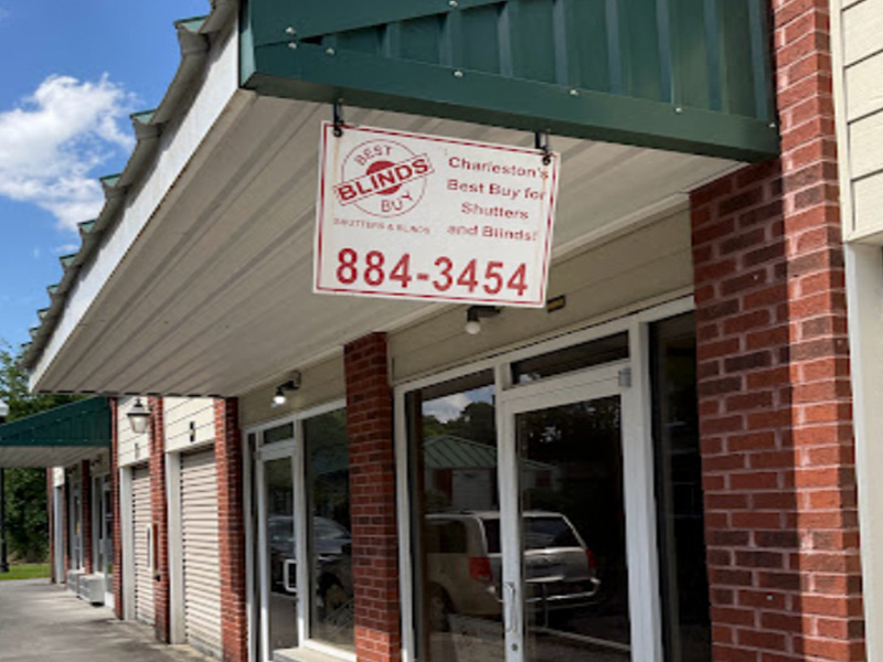 The image shows a sign for a business, specifically 'Blinds & Shutters,' with a phone number and an address provided. It is situated outside of a building with a brick facade under a clear sky.