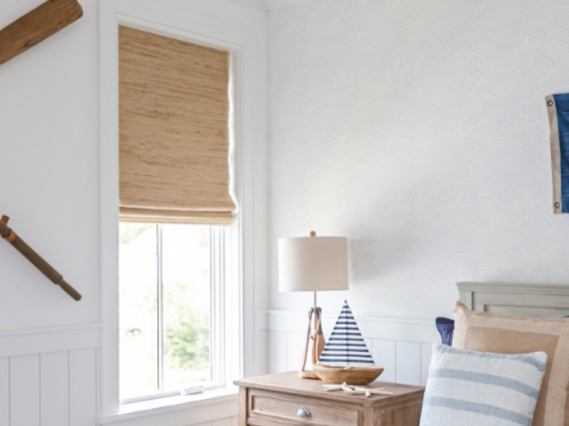 The image shows an interior room with a white wall, a window dressed with vertical blinds, and a blue and white flag hanging above the window. There is a wooden bed frame on the right side of the image with a pillow in the foreground. To the left, there s a small wooden nightstand with a lamp on it. A framed picture hangs on the wall to the left of the window, and a baseball bat leans against the wall above the nightstand. The room has a cozy and inviting atmosphere.