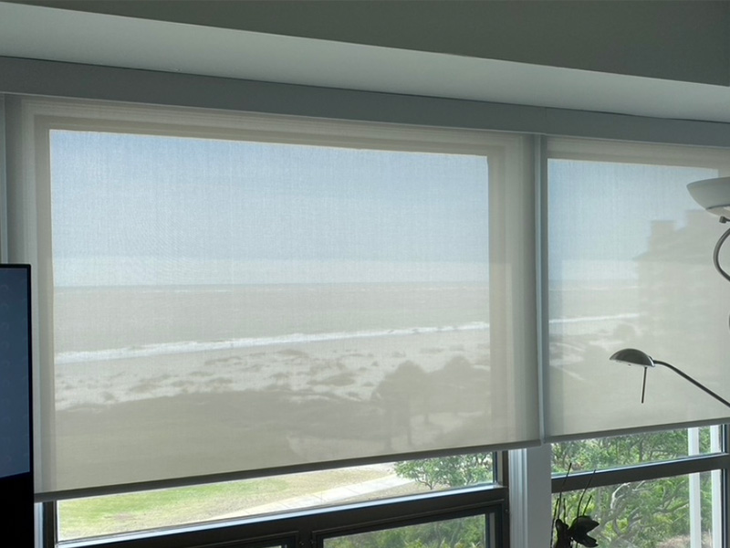 A room with a large window covered by black roller blinds, allowing a view of the ocean and sky.